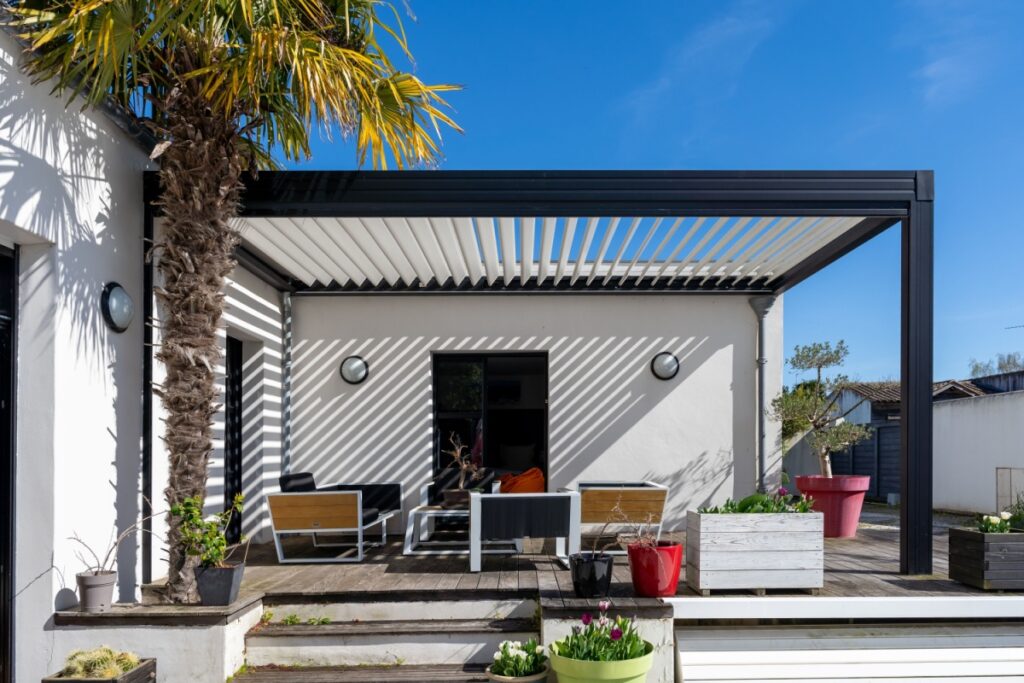 Moderna pergola bioclimatica addossata a una casa bianca, con lamelle orientabili che creano ombre geometriche su un patio in legno arredato