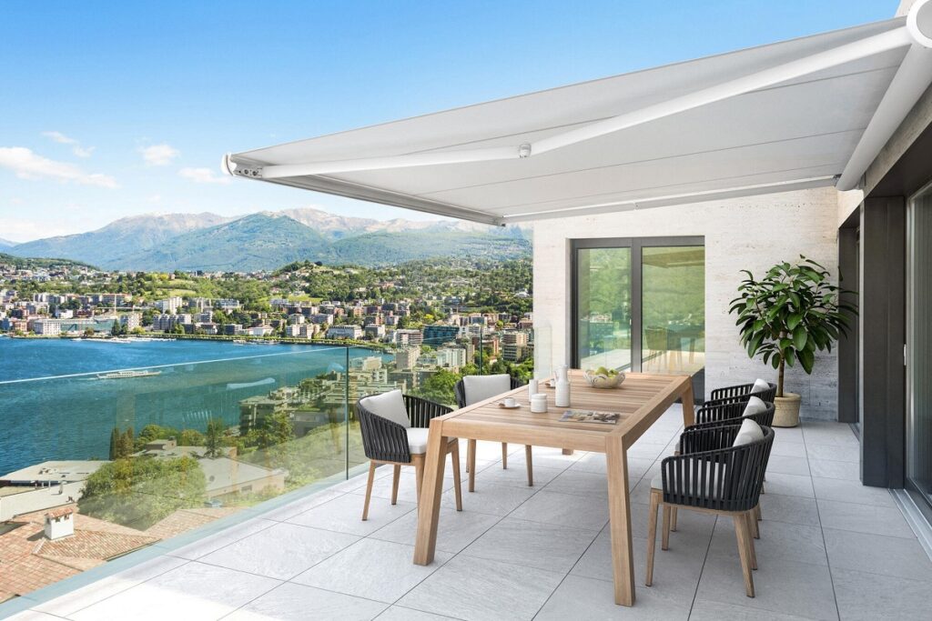 Ampio terrazzo panoramico con vista lago, arredato con zona pranzo e ombreggiato da una moderna tenda da sole bianca a bracci estensibili aperta.