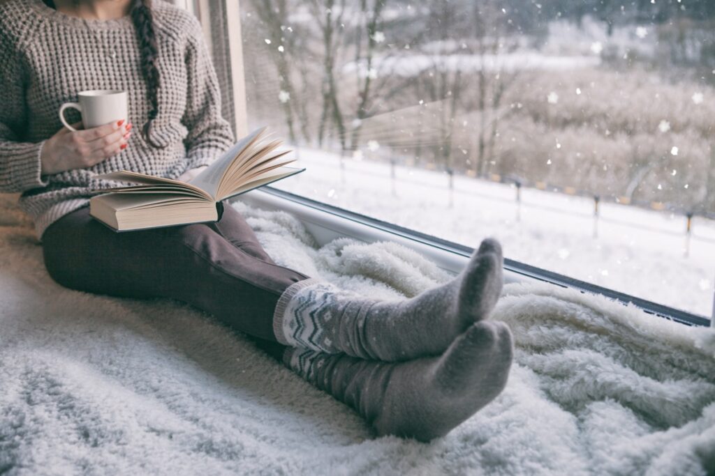 persona siede al caldo su una morbida coperta accanto a una finestra isolante, leggendo un libro e tenendo una tazza fumante mentre fuori nevica.