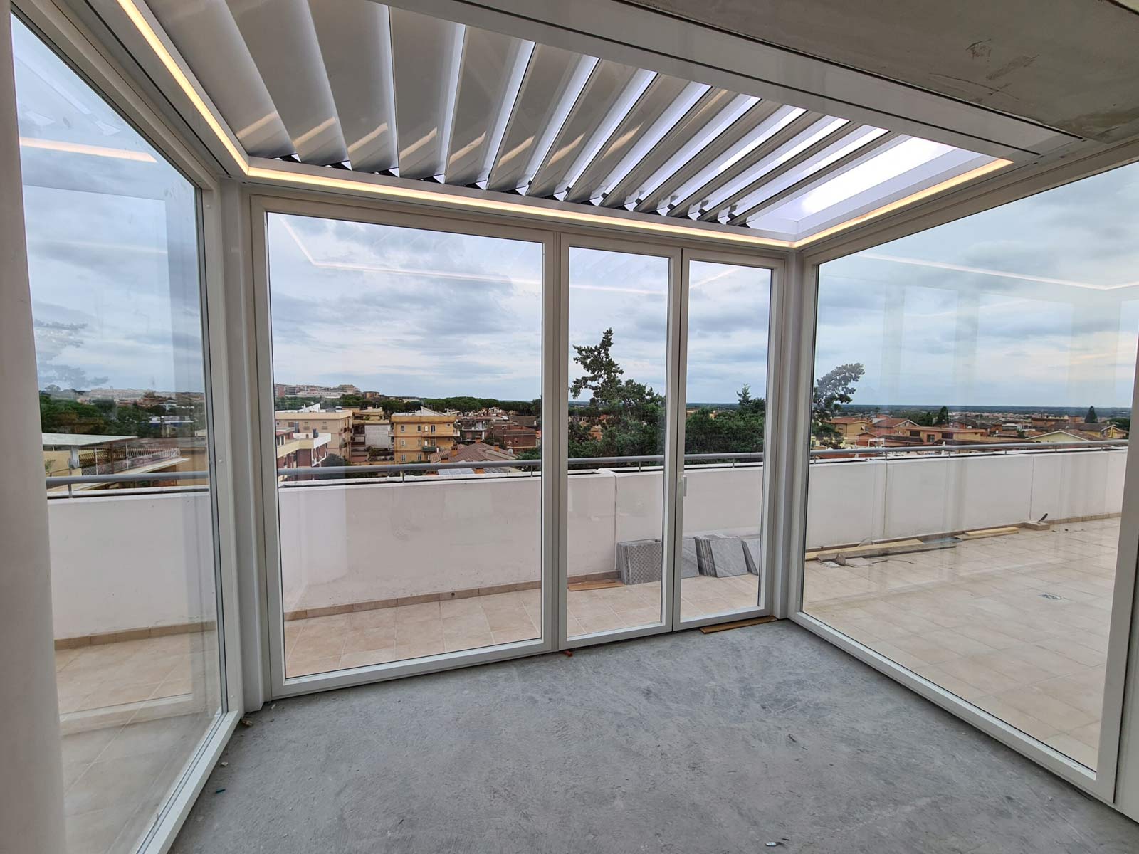 Interno di una pergola bioclimatica bianca con lame frangisole e vetrate scorrevoli su un attico a Roma